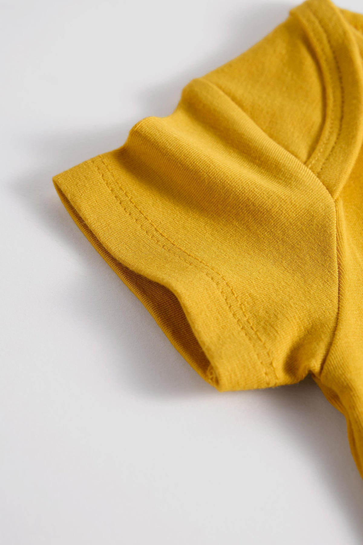 A close-up of a mustard yellow t-shirt sleeve with neat stitching, resting on a clean white surface.