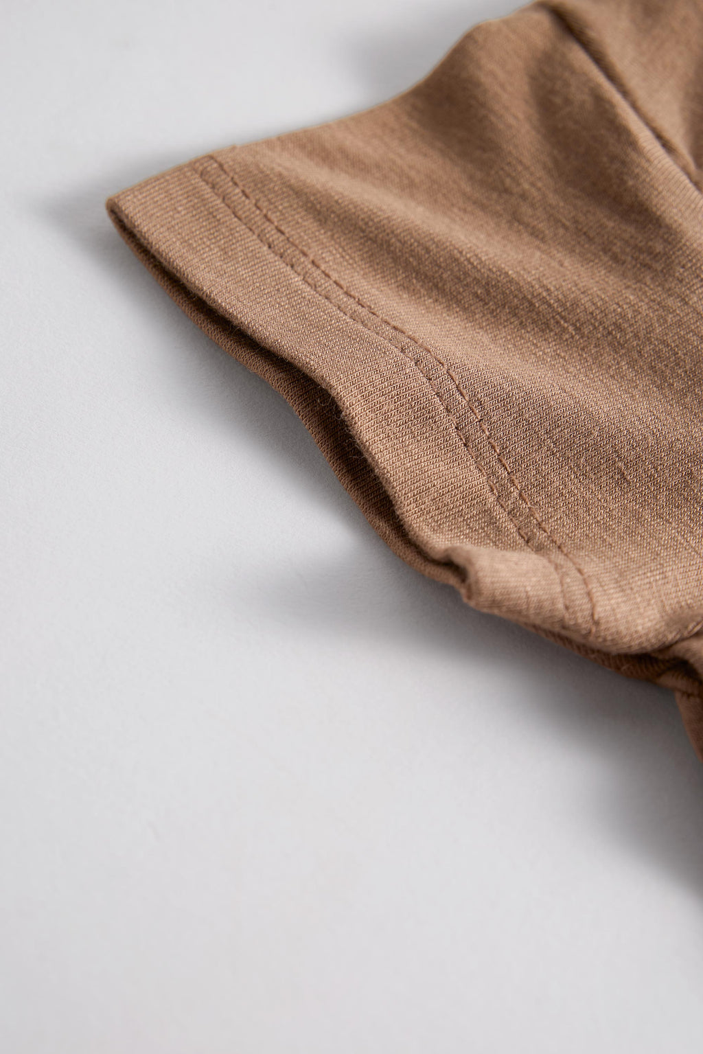 Close-up of a folded brown fabric garment, showing the texture and hem stitching on a light gray background.