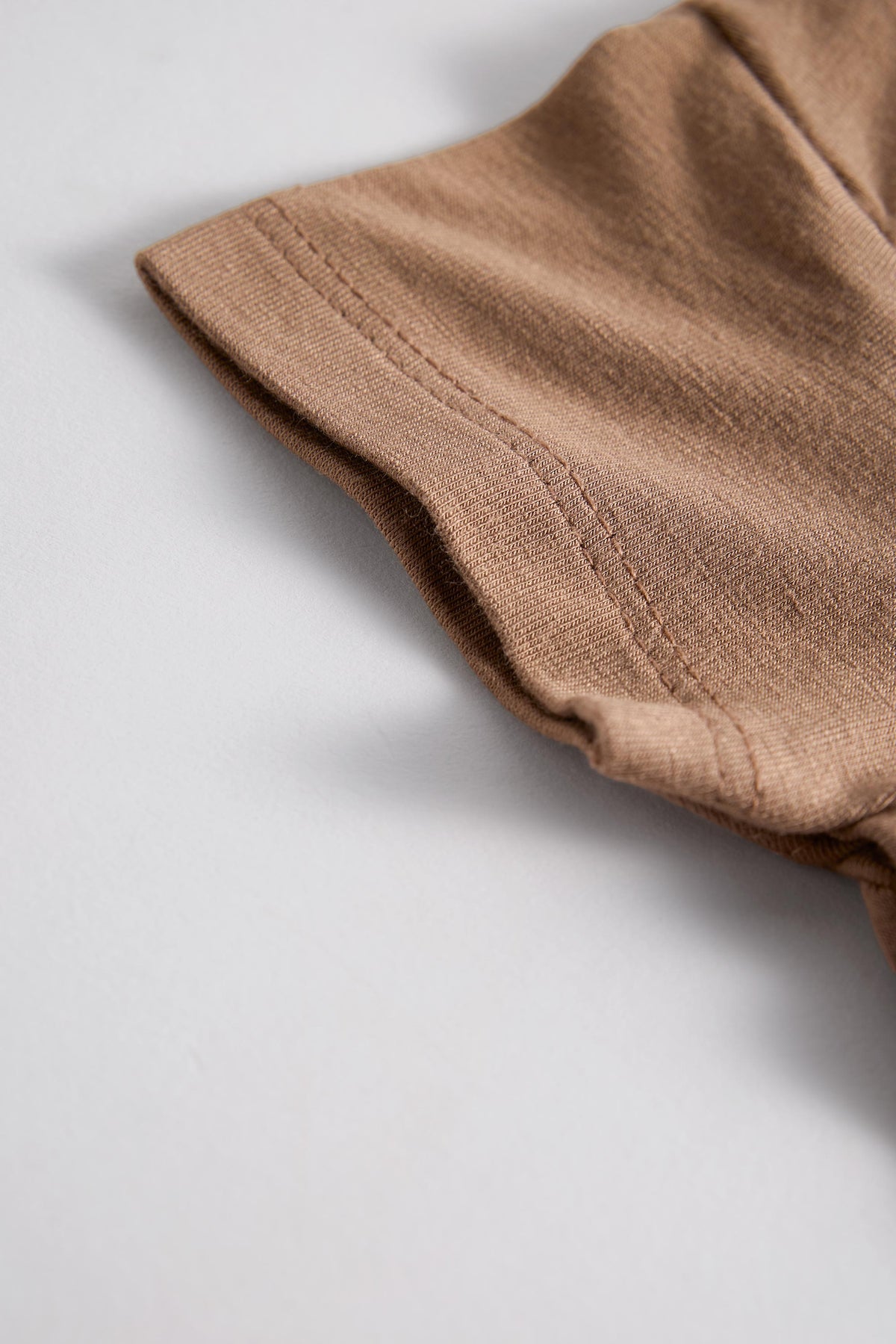 Close-up of a folded brown fabric garment, showing the texture and hem stitching on a light gray background.