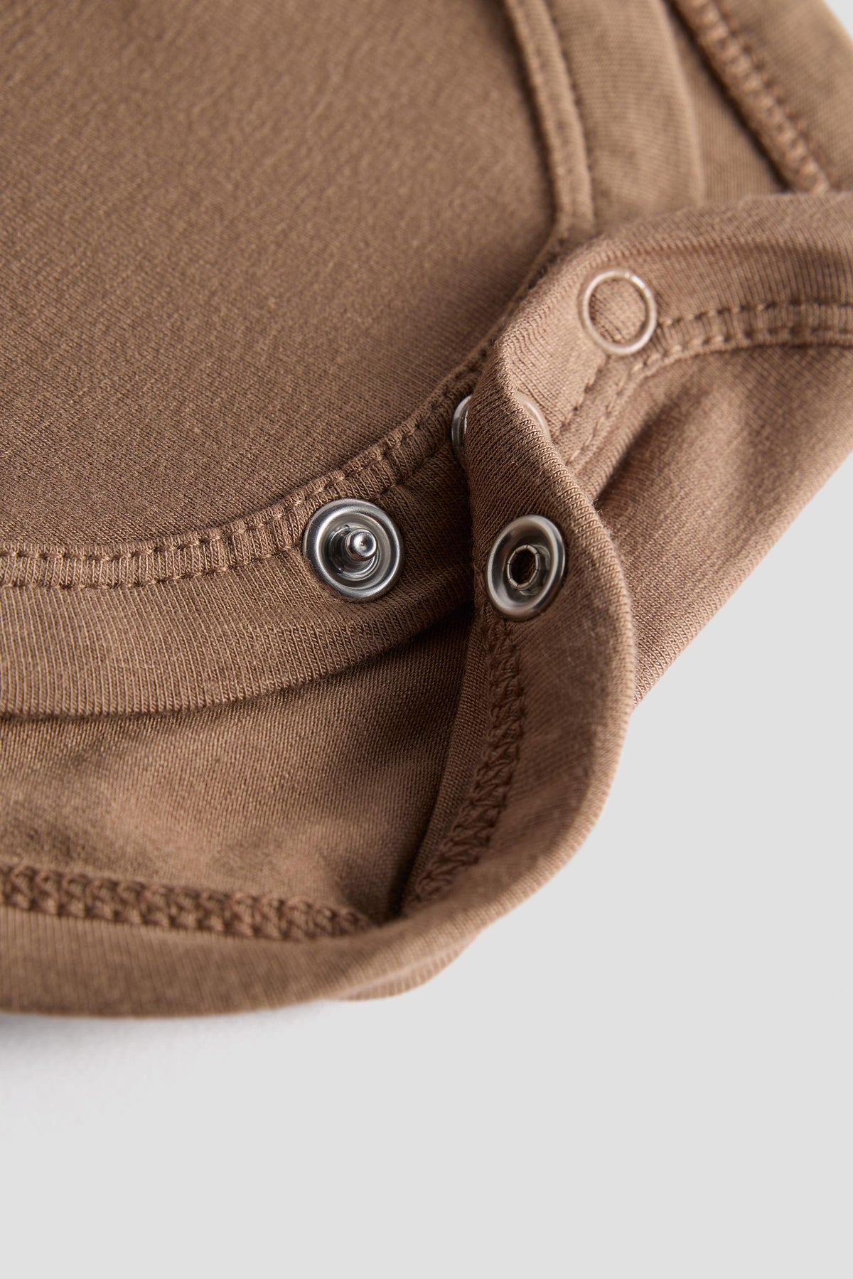 Close-up of a brown fabric garment with metal snap buttons and stitched edges on a light gray background.