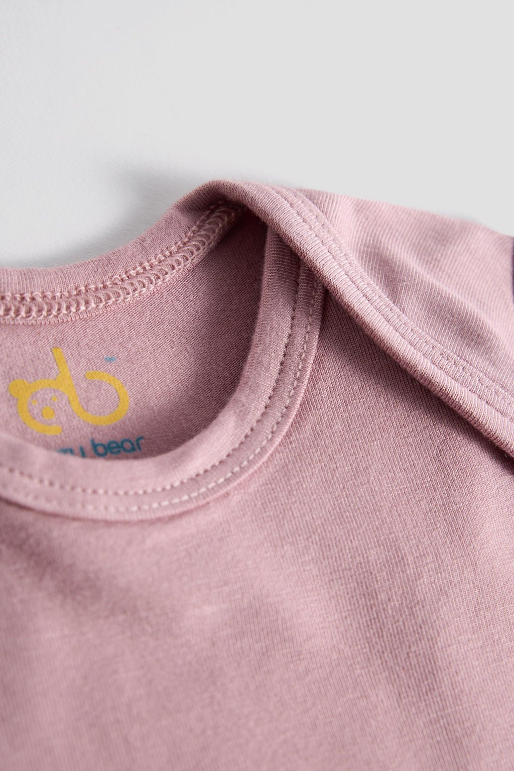 Close-up of a dusty pink baby bodysuit showing the inner neck label with a yellow and blue 'du bear' logo, set against a neutral light gray background.