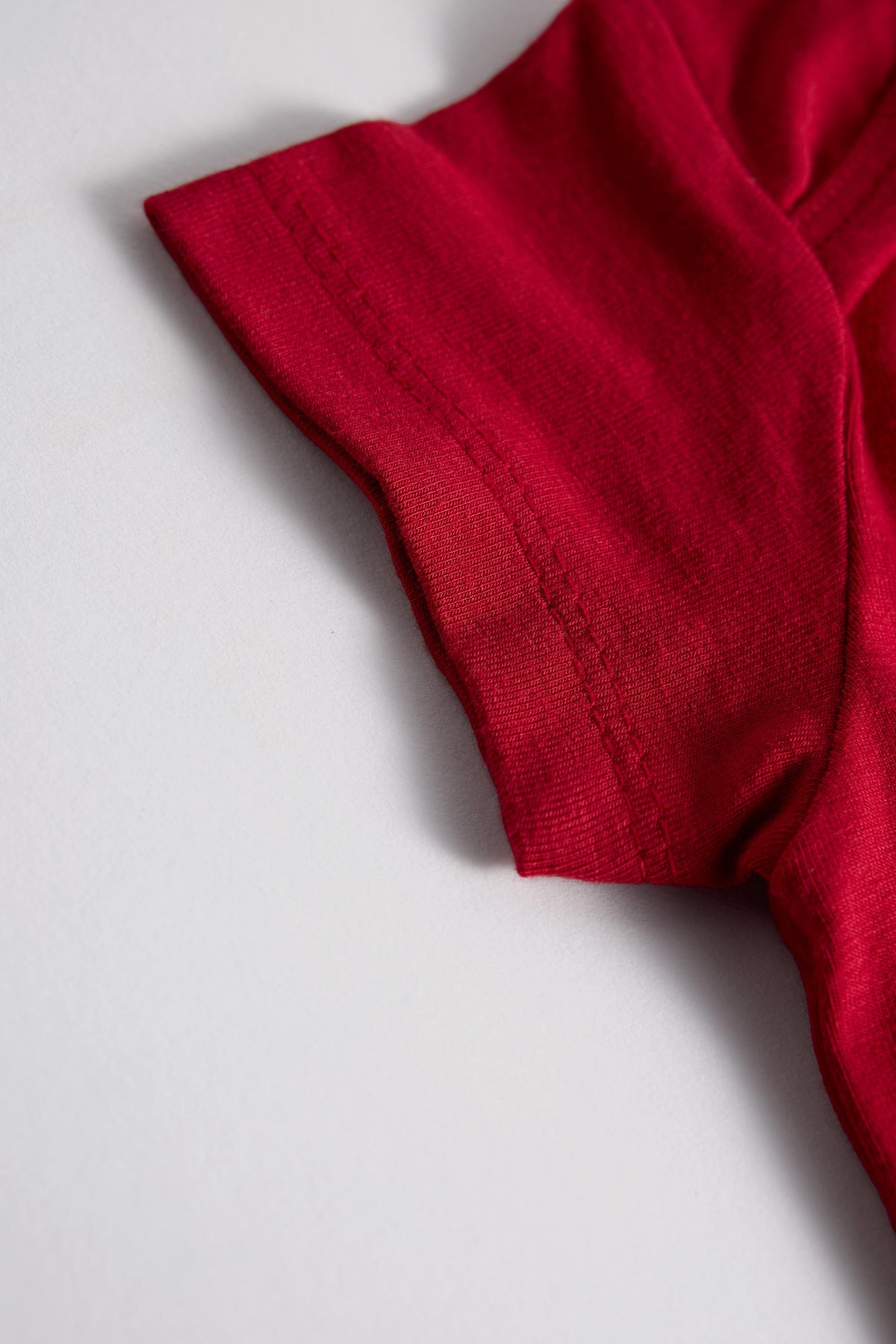 A close-up detail shot of a bright red short-sleeve garment, showing the ribbed knit fabric and sleeve cuff on a light gray background.