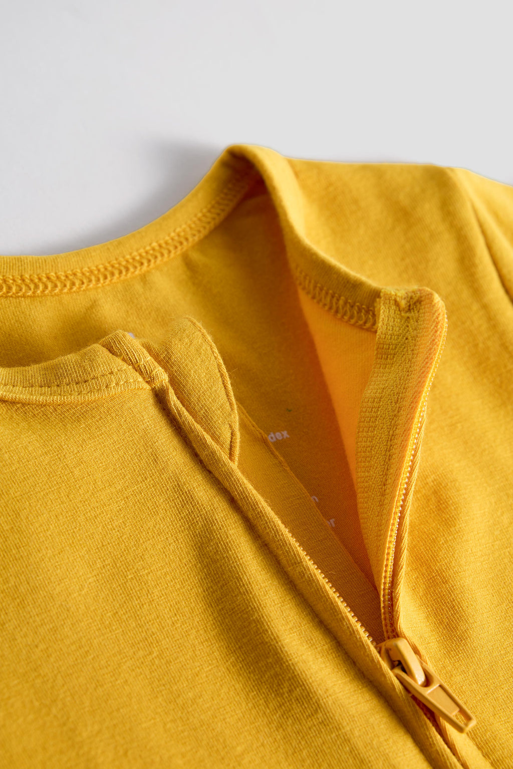 Close-up of a mustard yellow knit garment with a zipper neckline and ribbed trim, photographed against a light gray background.