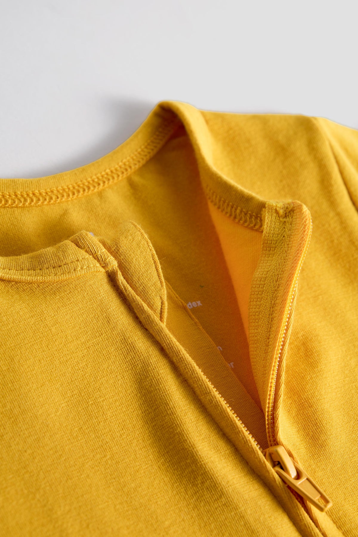 Close-up of a mustard yellow knit garment with a zipper neckline and ribbed trim, photographed against a light gray background.