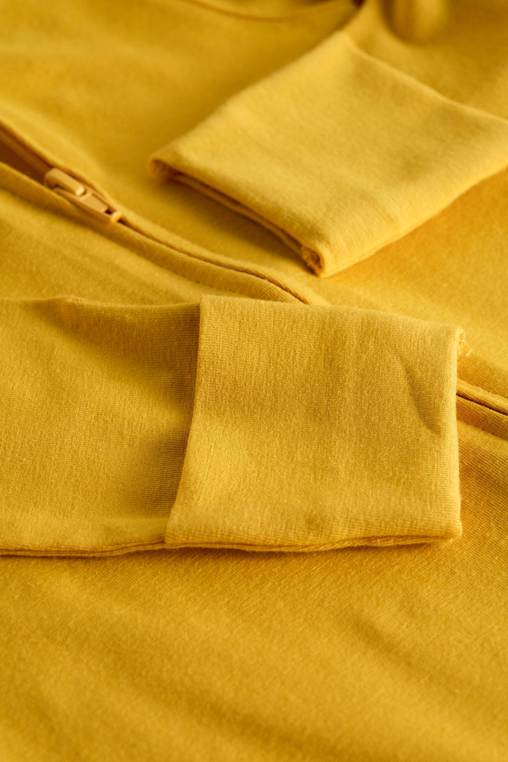 Close-up of a mustard yellow zip-up garment with folded cuffs, showing the soft fabric texture and zipper detail.