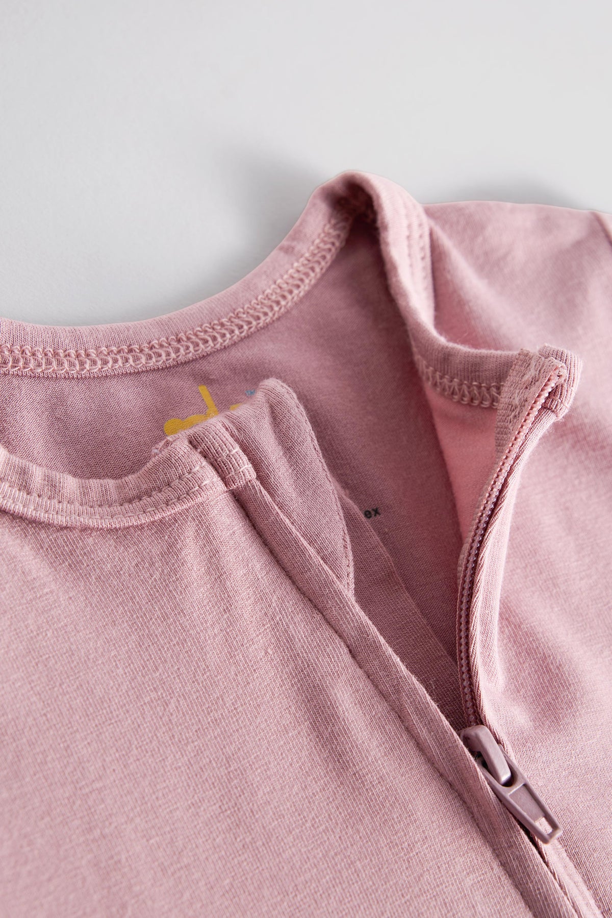 Close-up of a dusty pink zip-up baby bodysuit or long-sleeve top, showing the ribbed collar, zipper, and soft knit fabric texture against a light gray background.