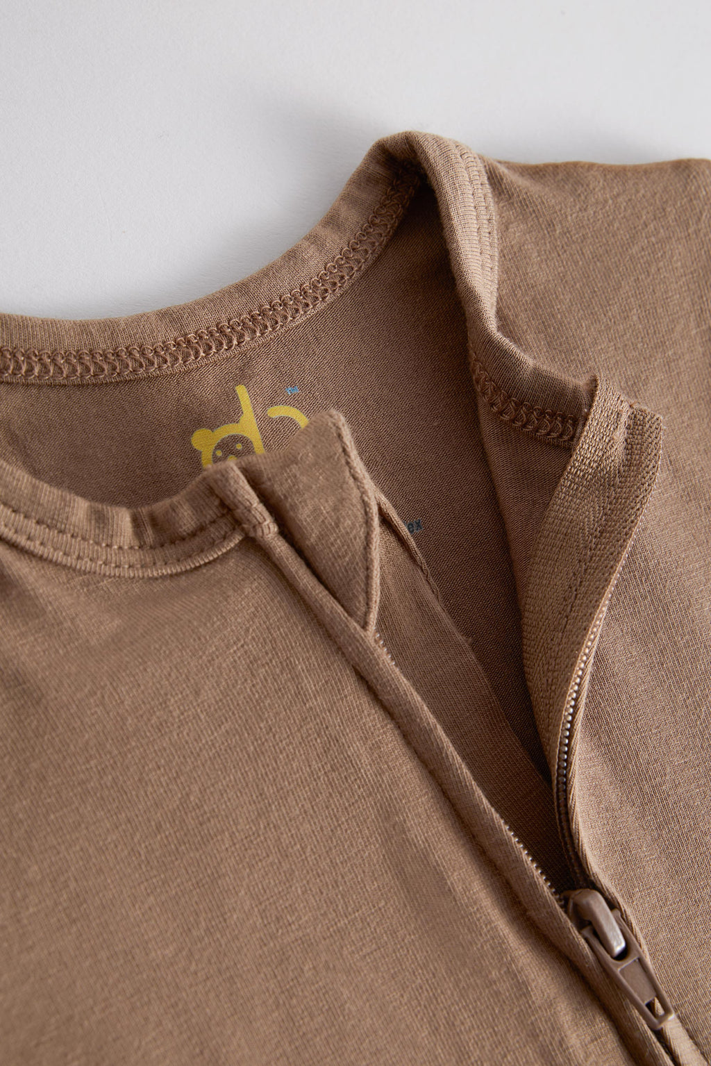 Close-up of a brown zippered garment, likely a hoodie or sweatshirt, showing the collar, ribbed neckline, zipper pull, and a small yellow graphic printed inside the collar.