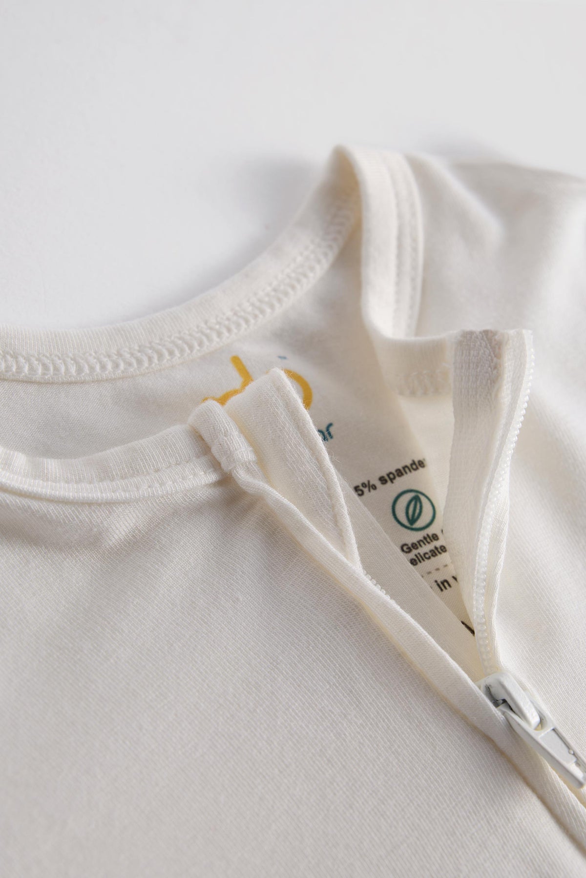 Close-up of a cream-colored zippered t-shirt showing the inner collar label with '5% spandex' and 'Gentle on skin' text.