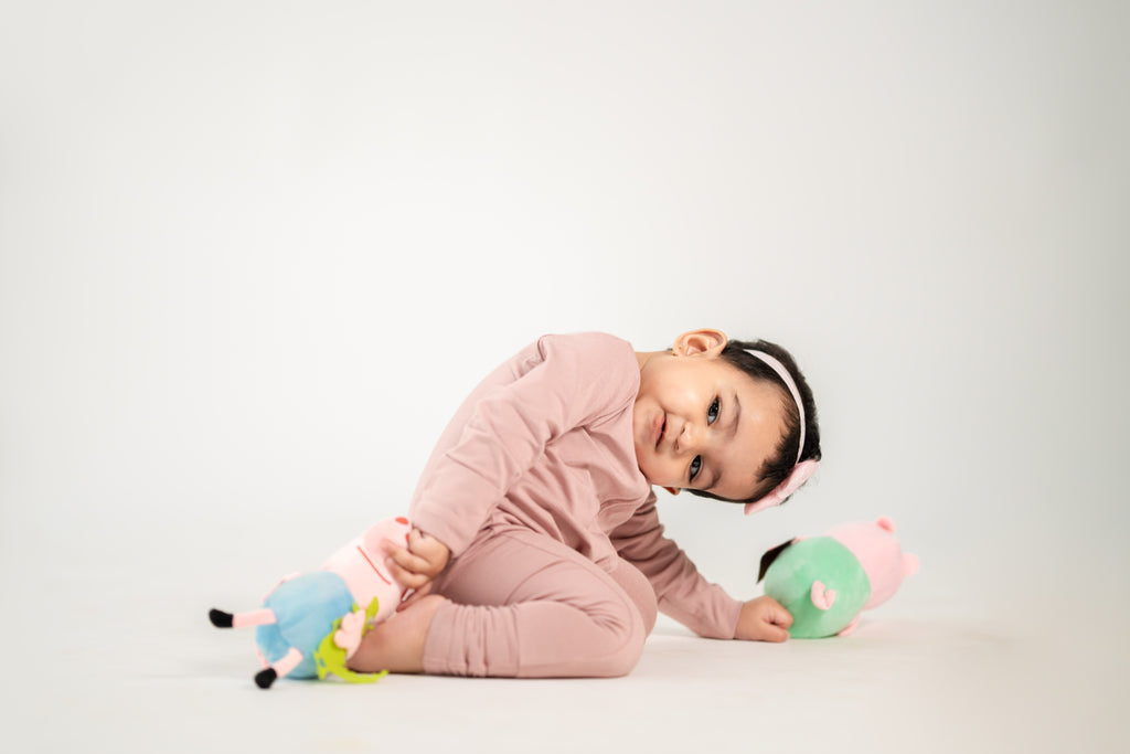 A young child wearing a pink long-sleeve pajama set and a pink headband lies on a light gray studio floor, holding a small plush toy.