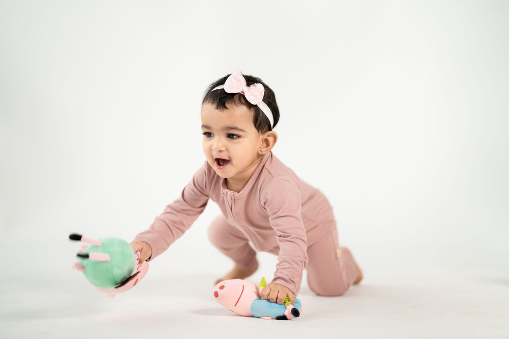 A baby girl crawling on a white studio floor, wearing a dusty pink long-sleeve jumpsuit and a pink bow headband, reaching for a green plush toy.