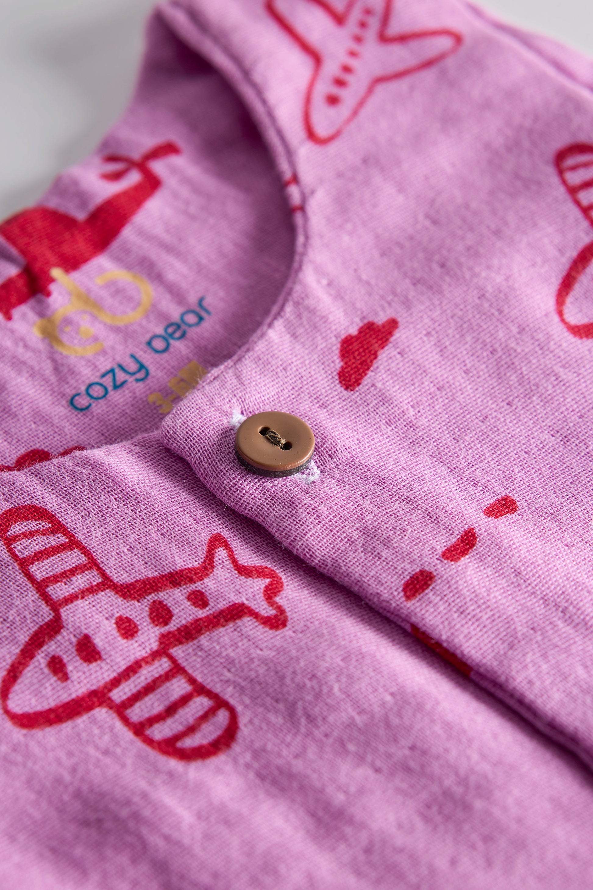 Close-up of pink fabric with red airplane patterns and a button, featuring the 'cozy bear' brand.