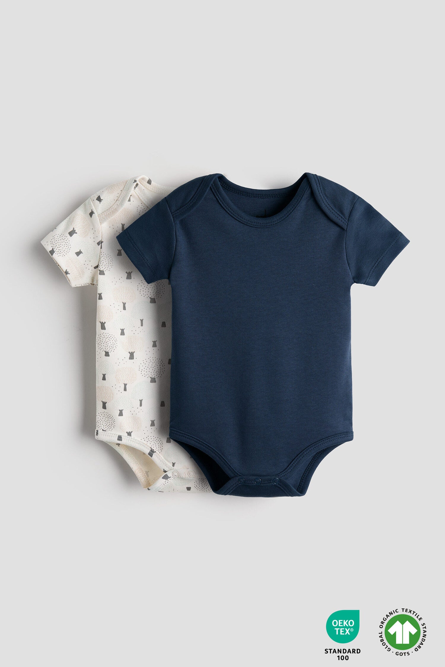 Navy blue baby bodysuit and patterned baby blanket on a light gray background with OEKO-TEX and Global Organic Textile Standards logos.