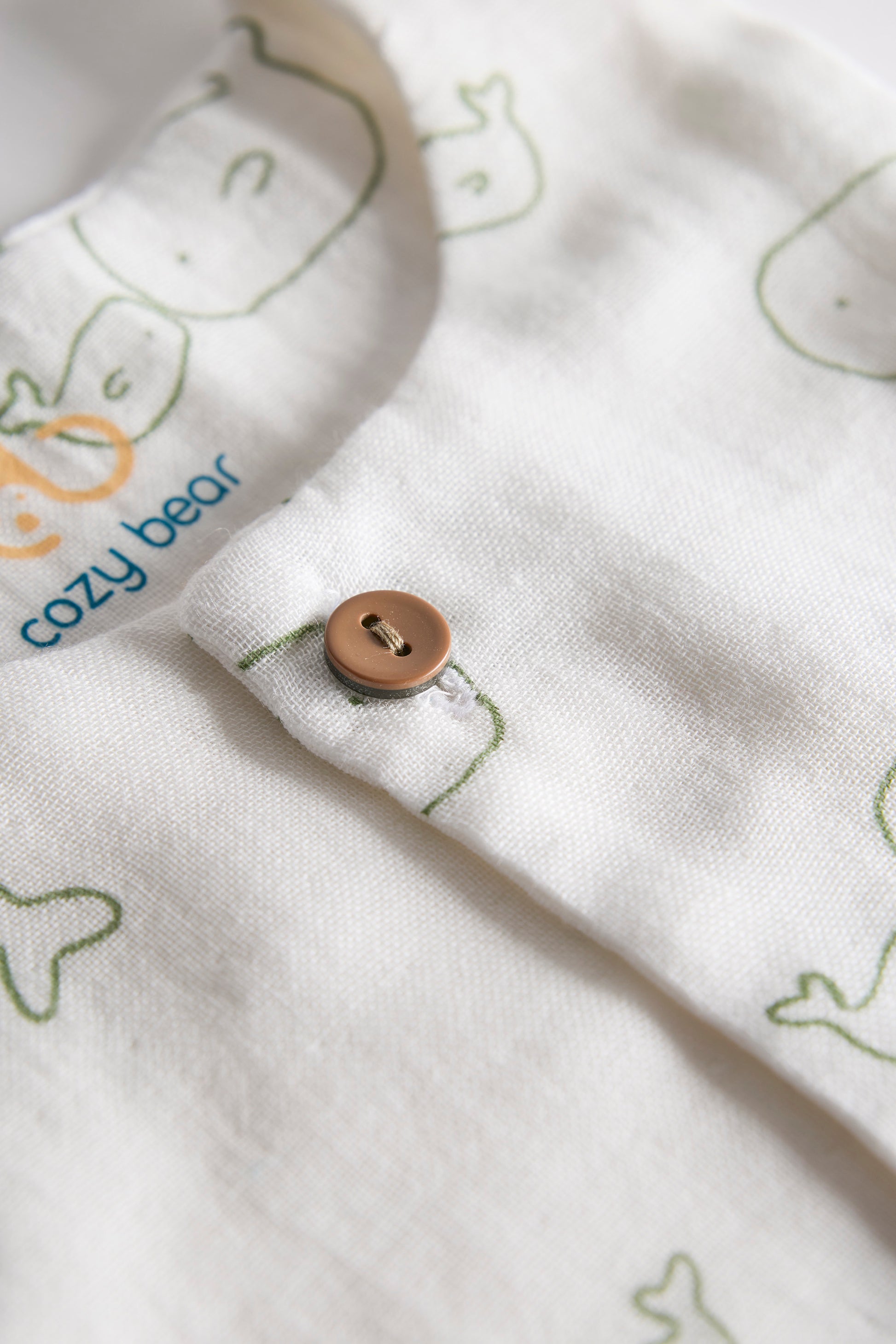Close-up of a white fabric with green bear prints and a brown button, featuring the 'cozy bear' brand.